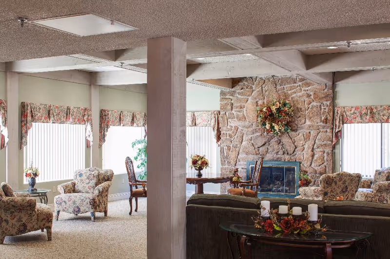 A cozy living room area in Mountain View Independent Living featuring floral upholstered armchairs, a stone fireplace decorated with a floral wreath, a round wooden table with chairs, and large windows with floral valances allowing natural light to fill the space.