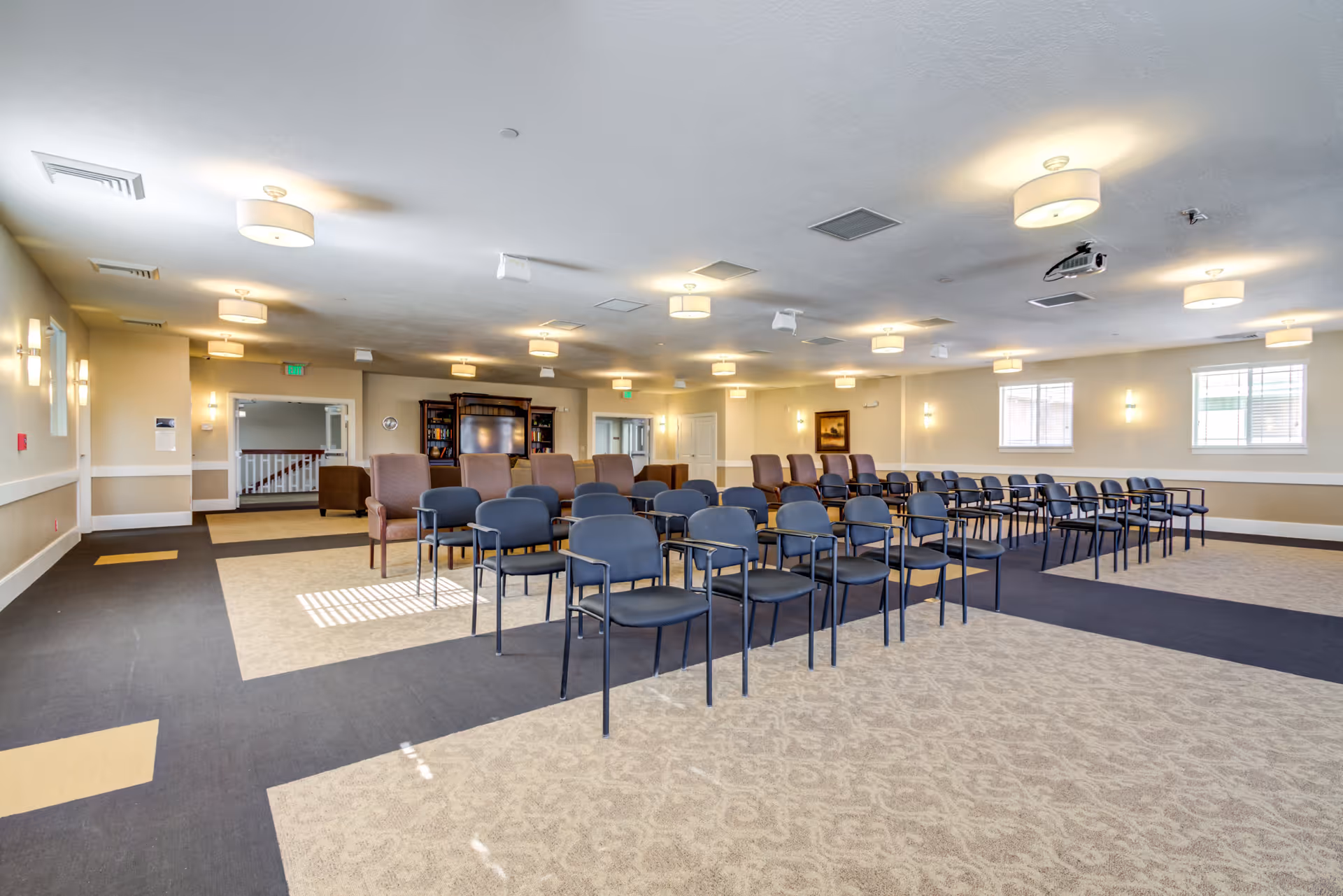 A spacious, well-lit meeting or activity room with rows of black chairs arranged facing a large TV and bookshelf unit. The room has beige walls, multiple ceiling lights, windows letting in natural light, and a patterned carpet with a dark walkway.