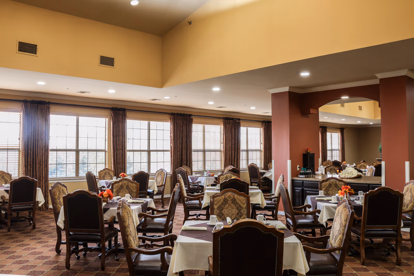 Spacious dining room in an assisted living facility with multiple set tables, upholstered chairs, and large windows along one wall.