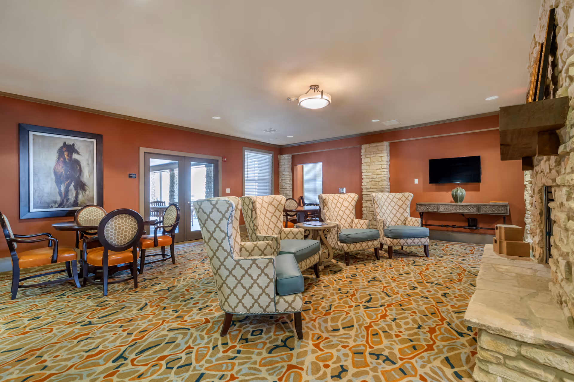 A spacious living room area with patterned carpet, four upholstered armchairs with blue cushions arranged around a small table, a stone fireplace on the right, a wall-mounted flat screen TV, and a wooden console table with a decorative vase. There are also two round tables with chairs near the back wall, a large painting of a horse on the left wall, and double glass doors leading outside.
