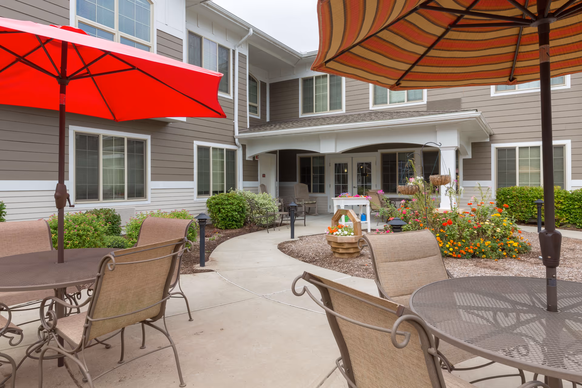 Courtyard patio with metal tables and chairs, colorful umbrellas, and flower beds outside a residential building.