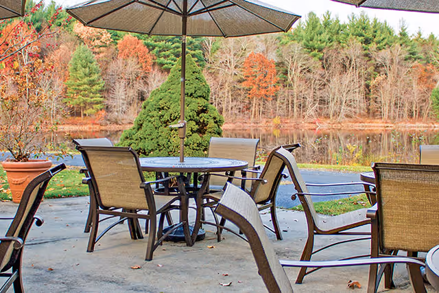Outdoor patio area with several metal and mesh chairs around round tables, each table shaded by a large umbrella. The patio overlooks a pond with trees in autumn colors in the background.