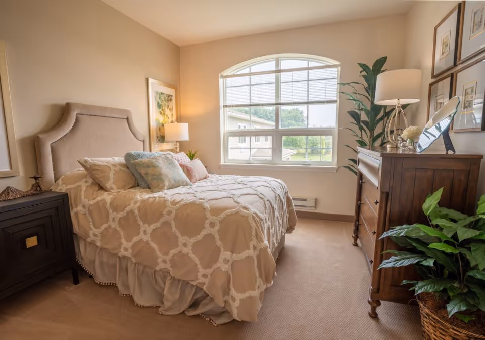 Sunlit bedroom with an upholstered bed, patterned bedding, nightstand and wooden dresser by a large arched window.