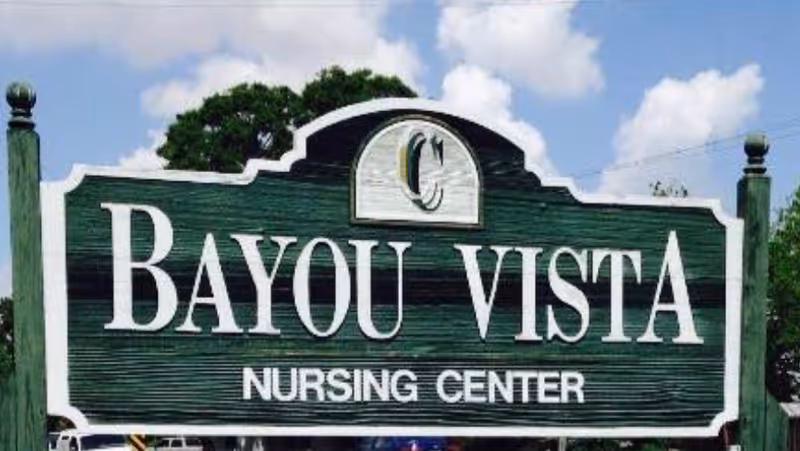 A green wooden sign with white text that reads 'Bayou Vista Nursing Center' against a partly cloudy blue sky background.
