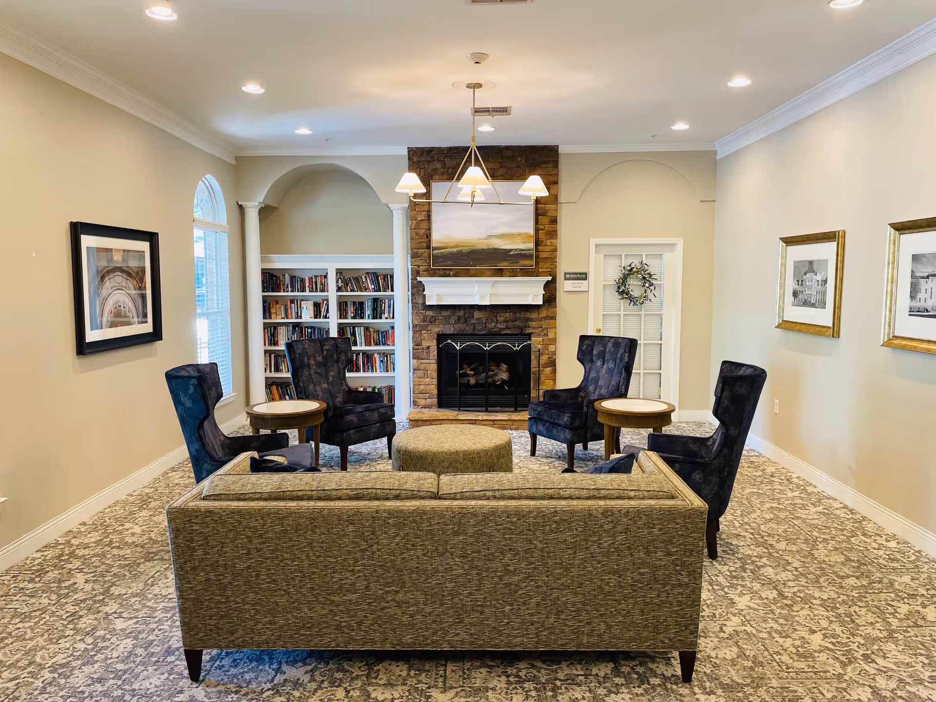 A cozy living room area with a beige couch facing a stone fireplace with a painting above it. There are four dark upholstered armchairs arranged around two small round wooden tables. Built-in bookshelves filled with books are on the left side of the fireplace. The room has light-colored walls with framed artwork and a patterned carpeted floor. Recessed ceiling lights and a chandelier provide lighting.