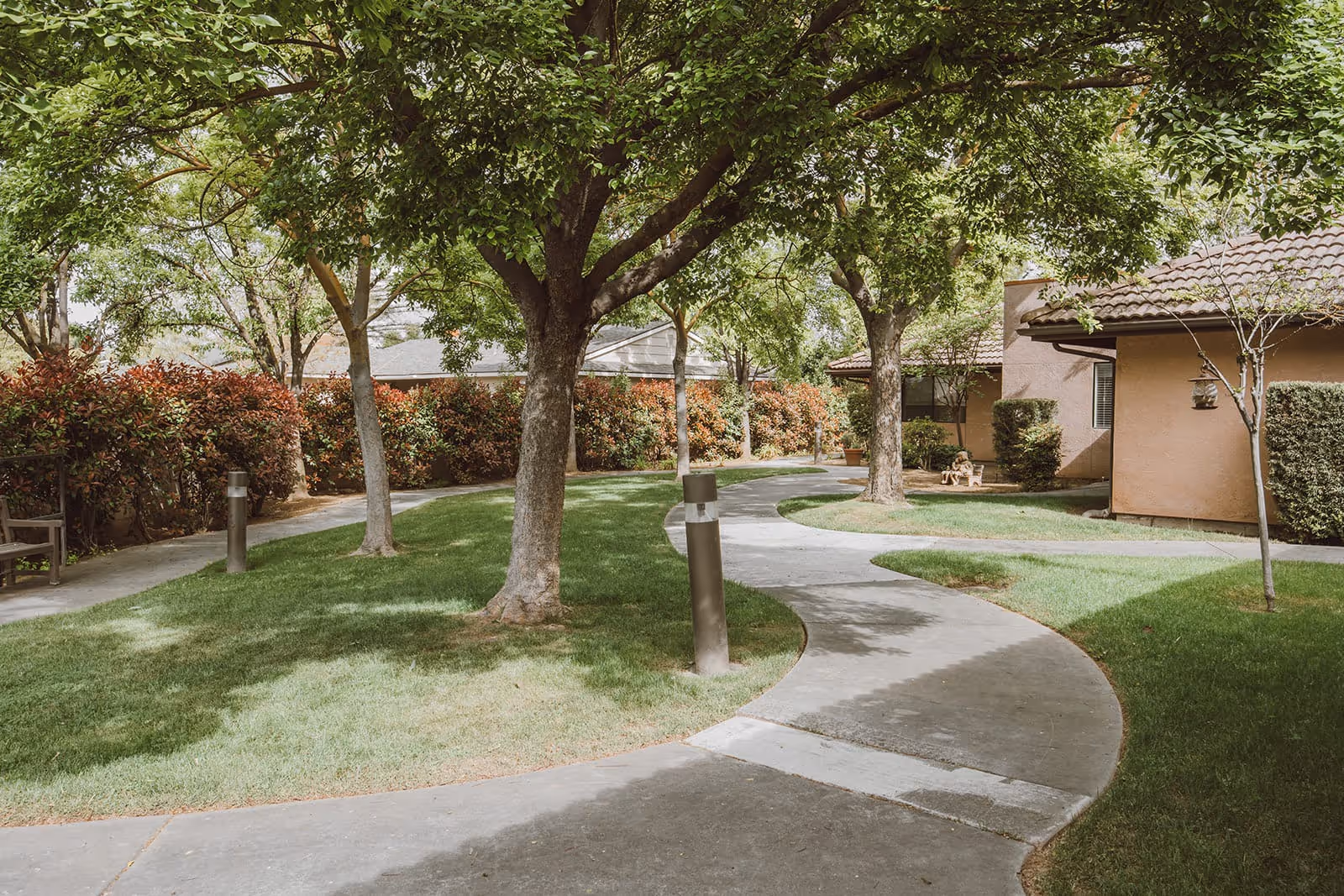 Curved concrete pathway winding through a green outdoor garden area with large trees, trimmed bushes, and a bench near a building with a tiled roof at Summerfield of Fresno.