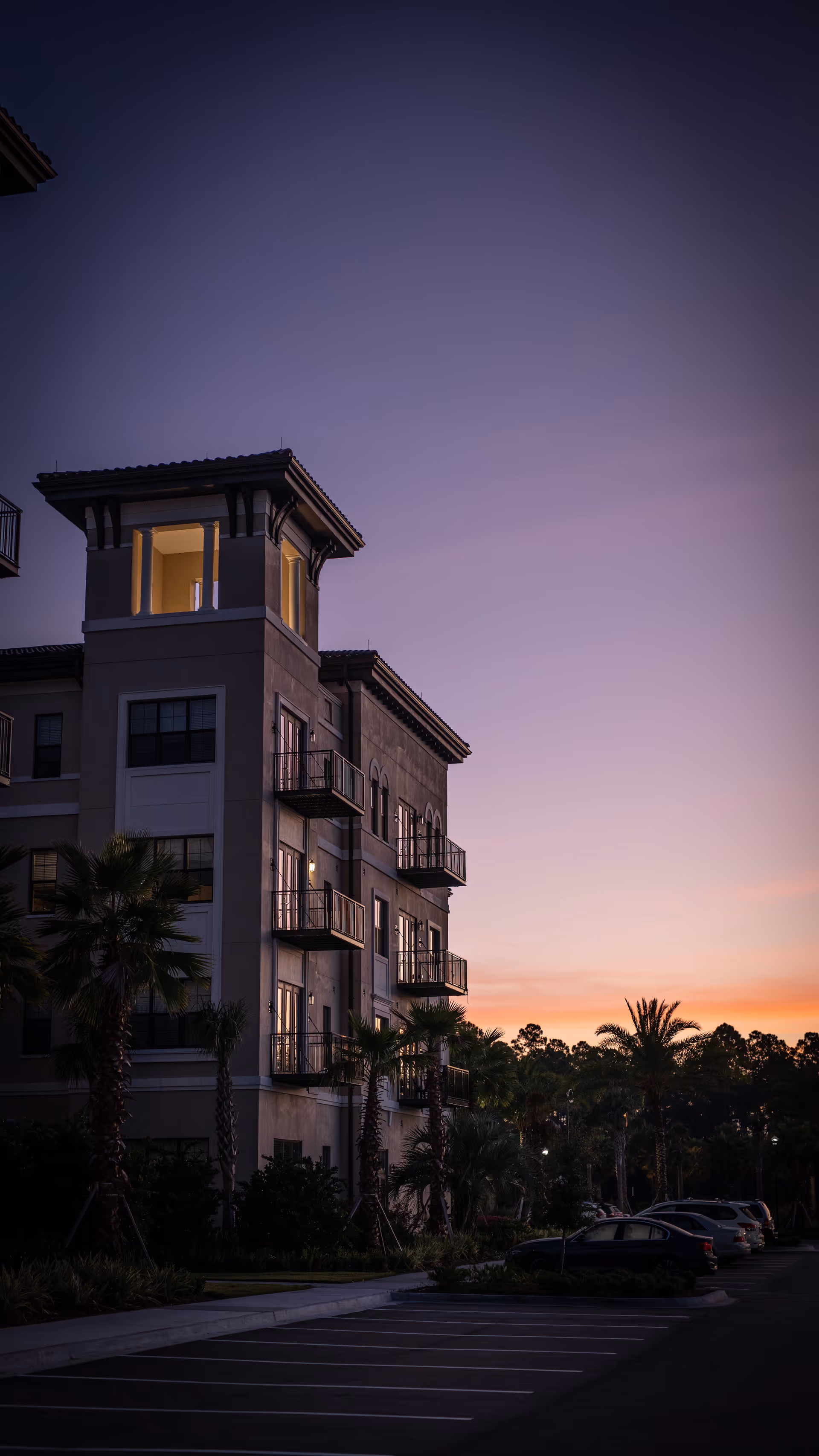 Exterior view of a multi-story residential building at dusk with balconies, palm trees, and parked cars in the foreground under a purple and orange sky.