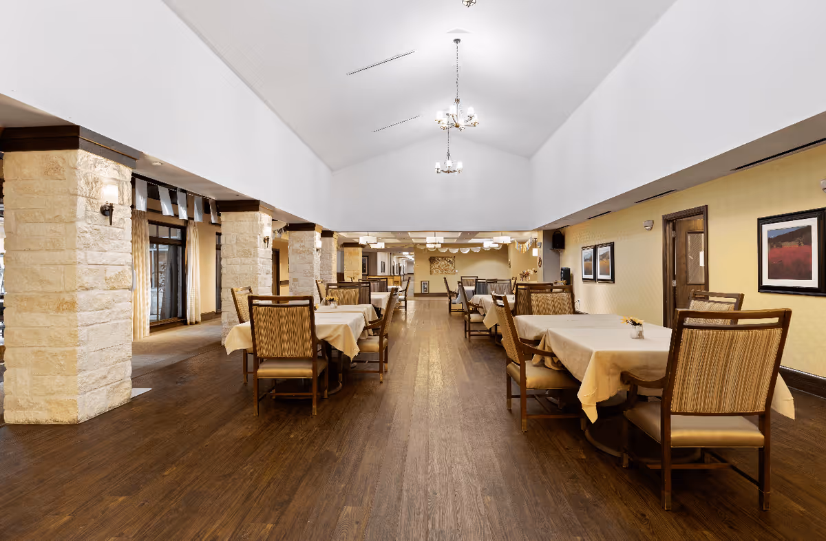 Spacious dining room with wooden floors, multiple tables covered with beige tablecloths, and cushioned chairs. Stone pillars line one side of the room, and the ceiling is vaulted with hanging chandeliers. The walls are decorated with framed artwork.