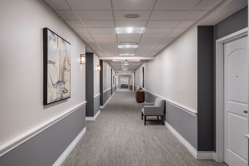A long, well-lit hallway in a senior living facility with gray and white walls, carpeted floor, modern wall sconces, framed abstract artwork, and a patterned armchair along the right side. There are doors on the right and decorative light fixtures on the ceiling.