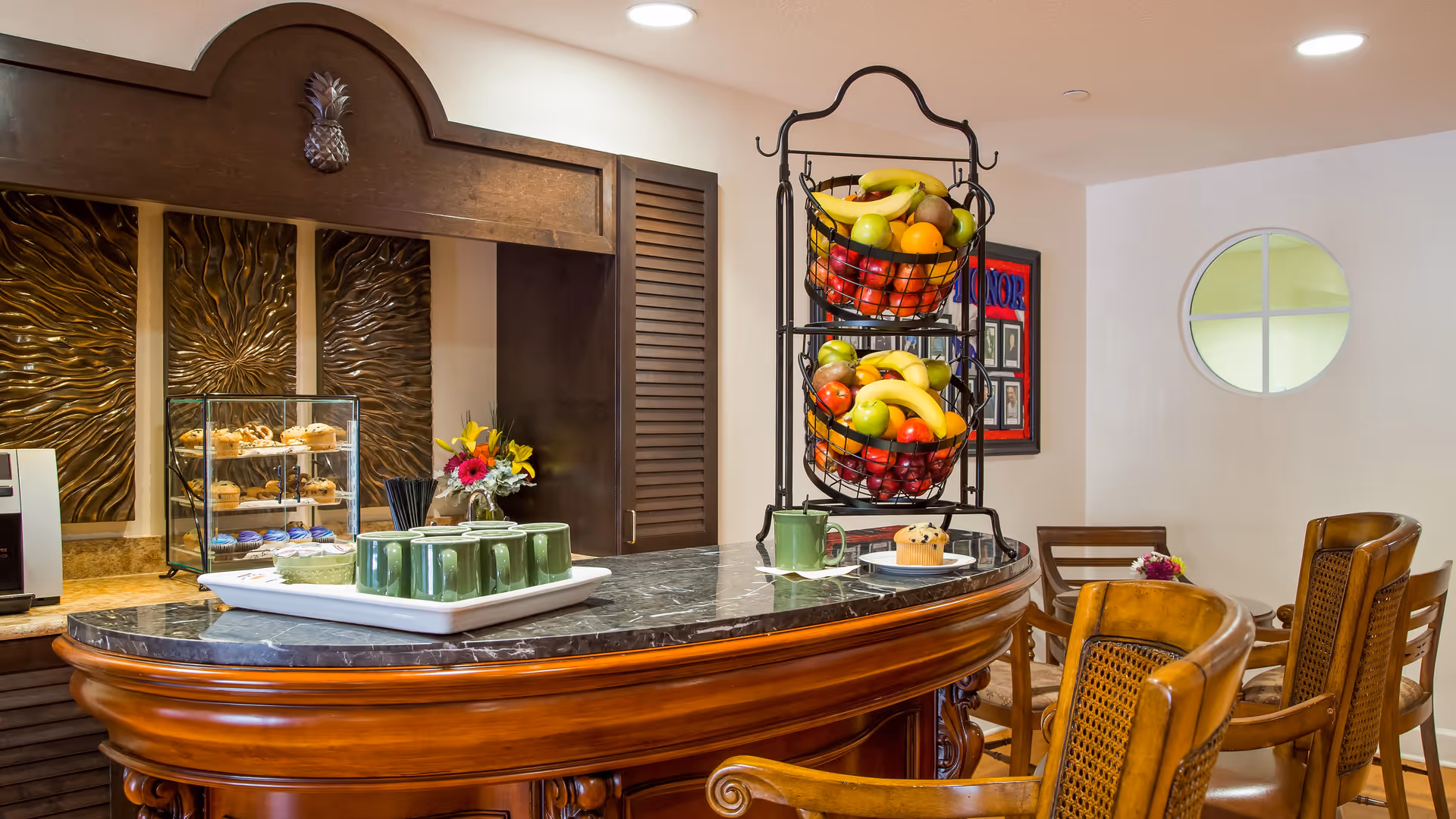 A cozy dining area with a curved wooden counter topped with a black marble surface. On the counter, there are green mugs on a white tray, a plate with a muffin, and a two-tiered fruit basket filled with bananas, apples, and oranges. Behind the counter, there is a display case with pastries and a decorative wall panel with a pineapple carving. Wooden chairs with woven backs are arranged around the counter, and a circular window is visible on the white wall.