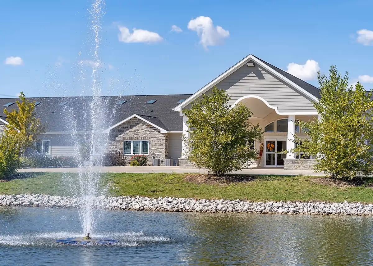 Front exterior of a single-story assisted living building with a covered entrance, landscaping, and a fountain in a pond in the foreground.