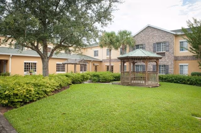 A grassy courtyard area with a wooden gazebo in the center, surrounded by bushes and trees. The courtyard is enclosed by a two-story building with beige and brick exterior walls and multiple windows.
