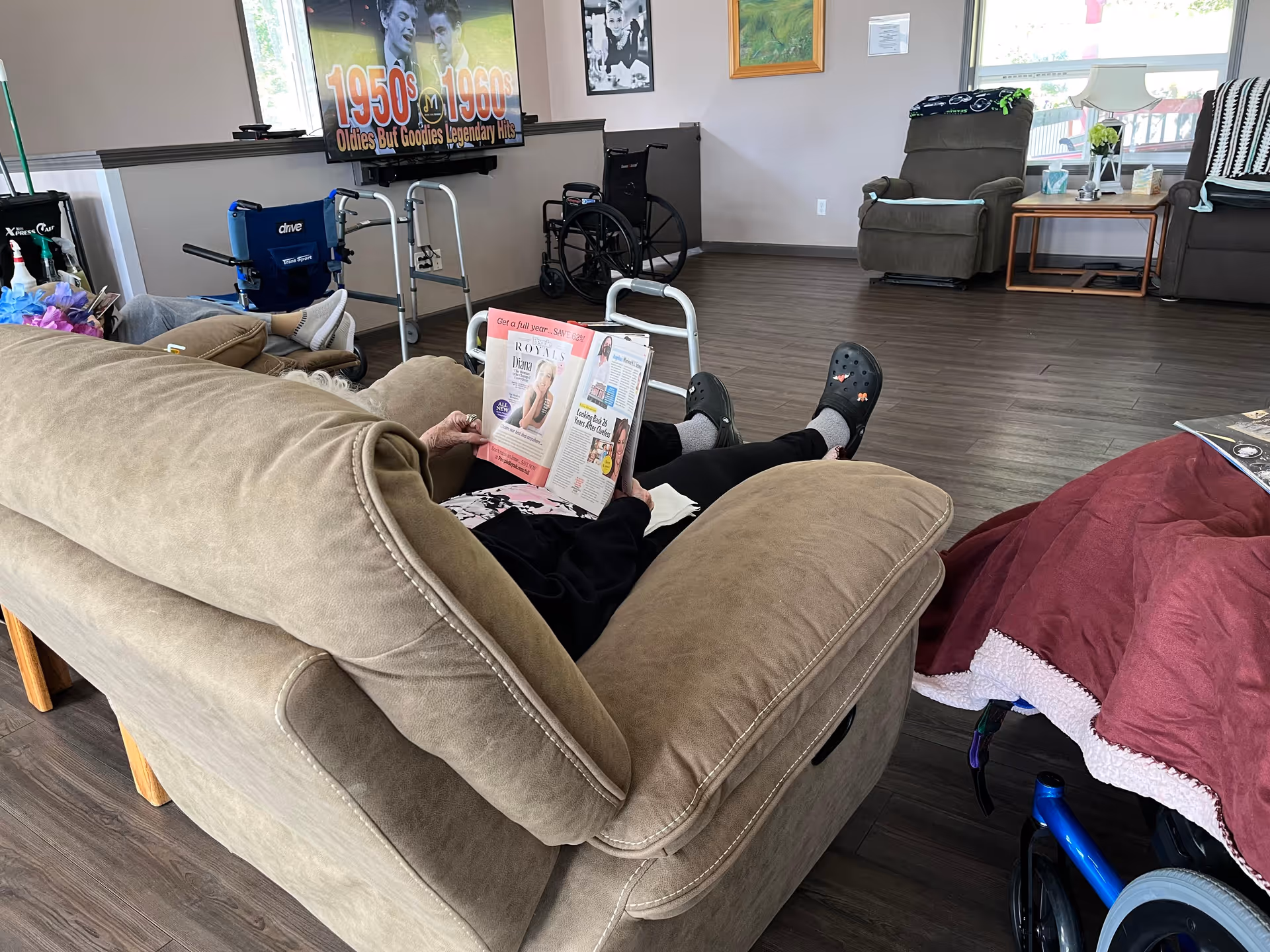 A common living area in a senior facility with a person reclining on a sofa reading a magazine, wheelchairs and a TV in the background.