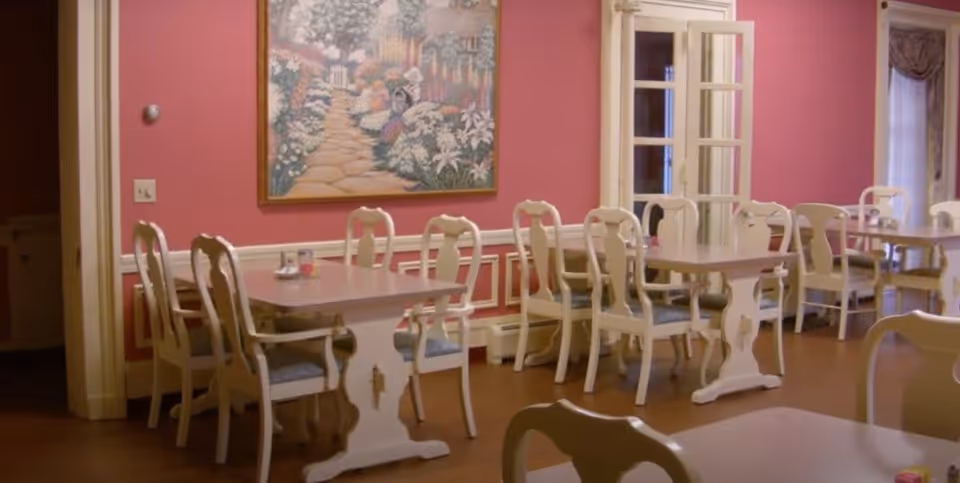 Dining room with multiple tables and white chairs with blue cushions. The walls are painted pink with white trim, and there is a large framed painting of a garden pathway on the wall. French doors and windows with curtains are visible in the background.