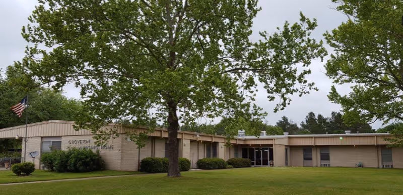 Exterior view of Groveton Nursing Home, a single-story building with beige brick walls, surrounded by green grass and trees. An American flag is visible on a flagpole near the building entrance.
