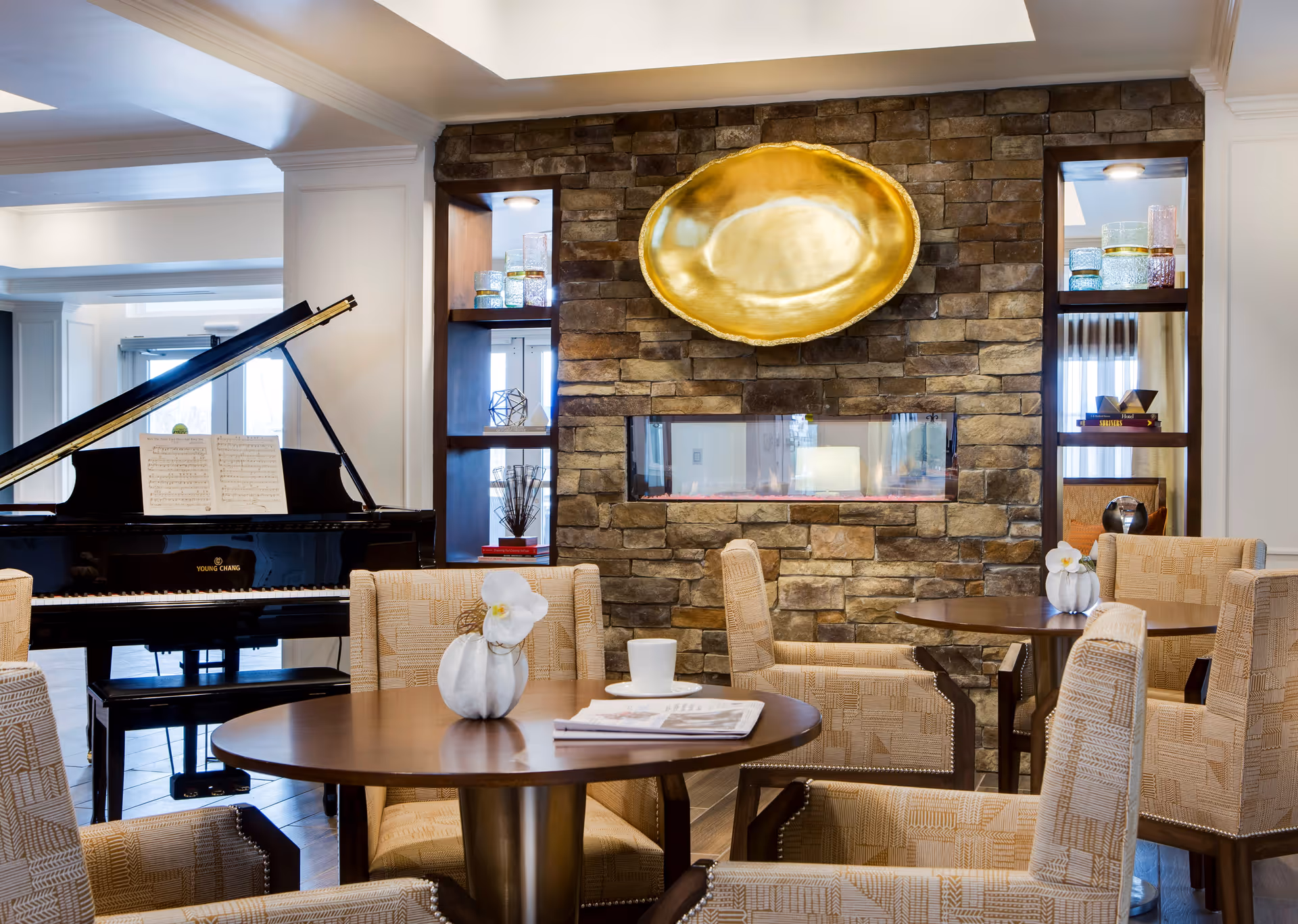 A cozy lounge area featuring a round wooden table surrounded by beige upholstered chairs. On the table is a small white pumpkin decoration and a newspaper. Behind the seating area is a stone wall with a modern rectangular fireplace and a large golden decorative plate mounted above it. Built-in shelves on either side of the fireplace hold decorative glassware and books. To the left, a black grand piano with sheet music is visible near a window.