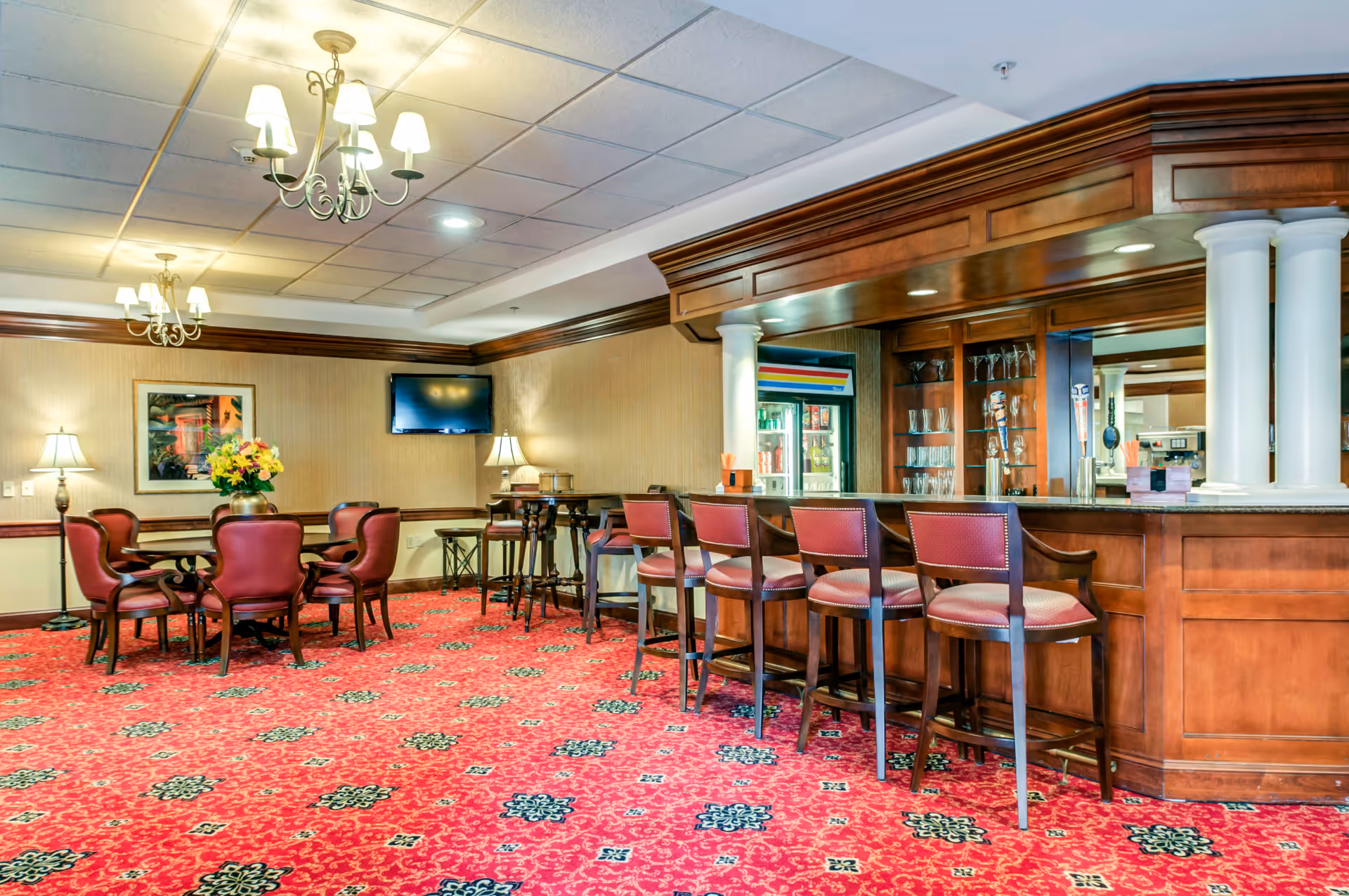 Carpeted lounge area with a wooden bar and barstools, a round table with chairs, chandeliers, and a wall-mounted TV.