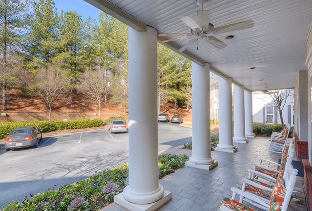 View of a covered porch with white columns and several rocking chairs with floral cushions, overlooking a parking lot with cars and trees in the background.