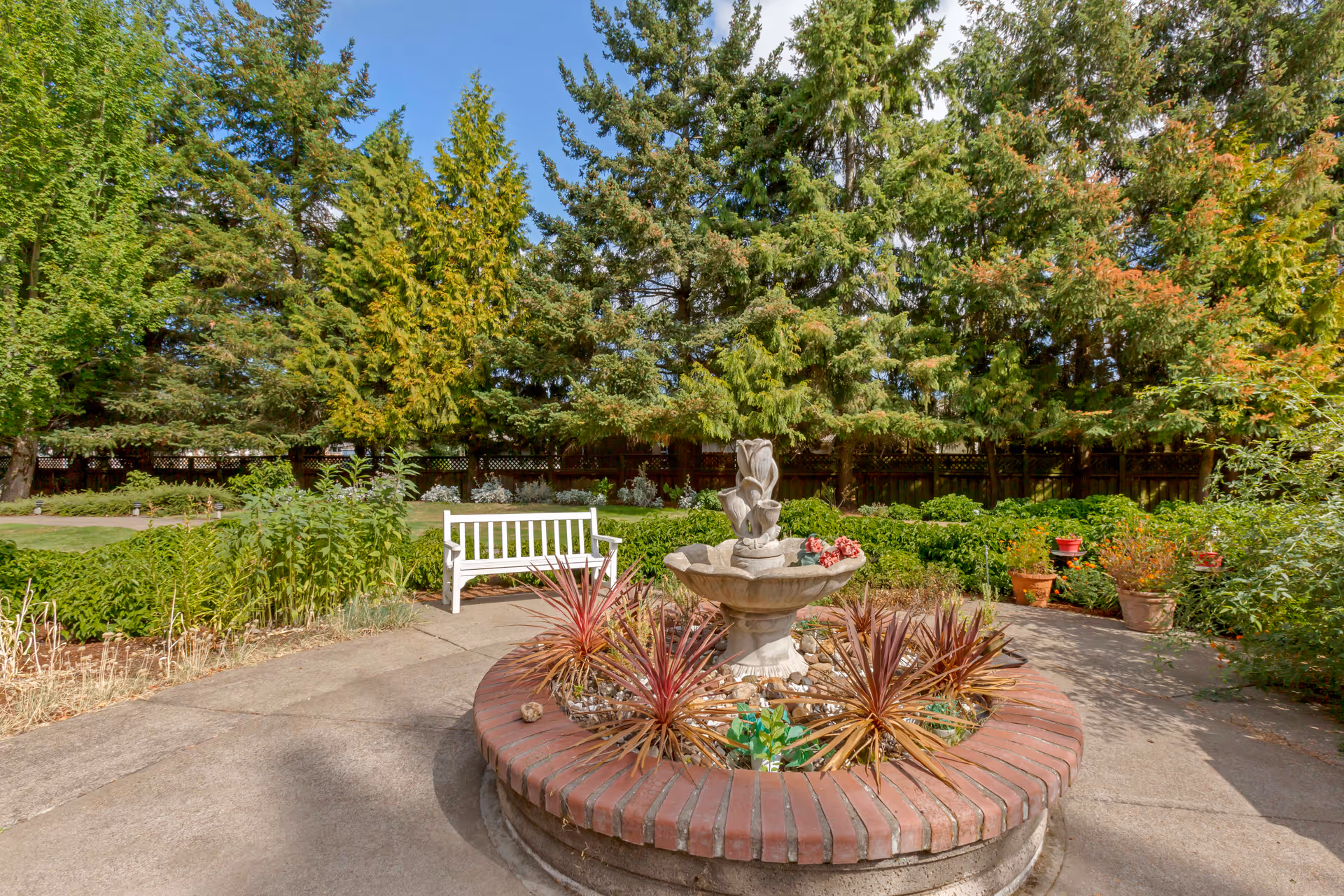 A peaceful outdoor garden area with a circular brick planter containing a decorative stone fountain and various plants. There is a white wooden bench nearby, surrounded by lush greenery and tall trees under a clear blue sky.