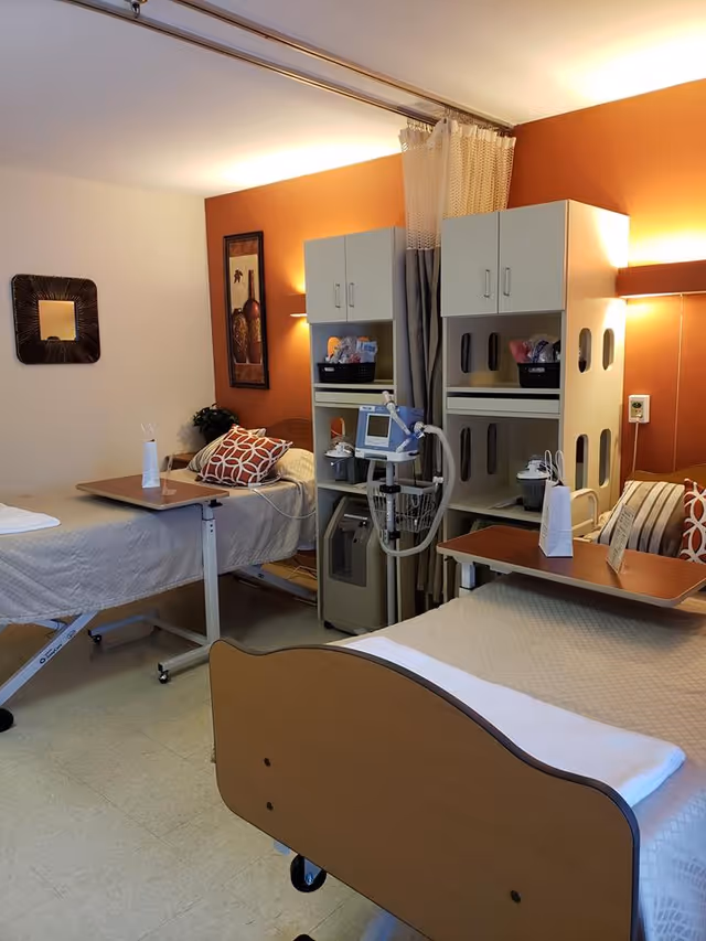 A shared patient room in a senior living facility with two hospital beds, each equipped with overbed tables and pillows. Between the beds are medical equipment and storage cabinets. The walls are painted in warm tones with decorative artwork and a mirror. A privacy curtain is partially drawn between the beds.