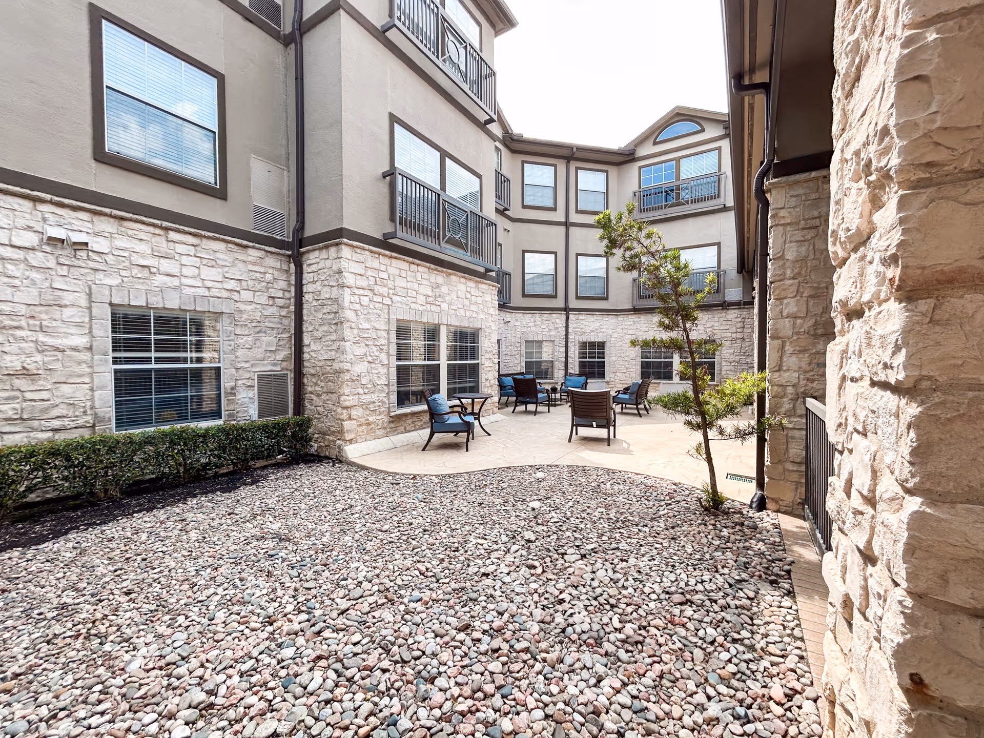 Outdoor courtyard area of a senior living facility with stone walls and multiple windows. The courtyard has a paved seating area with several chairs and small tables arranged for relaxation. There is a small tree and some shrubbery along the edges of the courtyard.