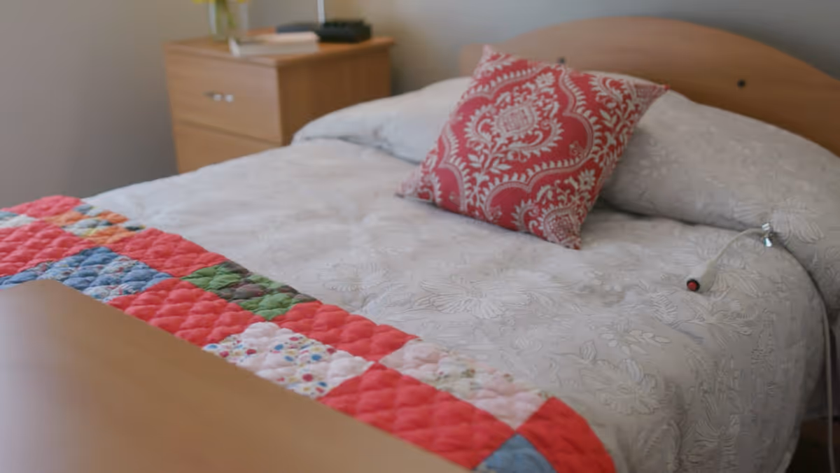 A neatly made bed with a white floral patterned bedspread, a colorful patchwork quilt at the foot, and a red decorative pillow with a white intricate design. Next to the bed is a wooden nightstand with a lamp and some items on top.
