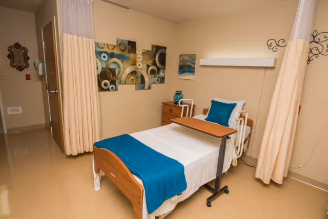 A tidy single patient room with a hospital-style bed, overbed table, teal accents, a bedside drawer, and privacy curtains.