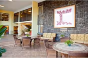 Indoor seating area with wicker chairs and tables, cushioned sofas against a stone wall, and a large framed artwork featuring a letter 'L' on the wall. The space has high ceilings and large windows, creating a bright and welcoming atmosphere.