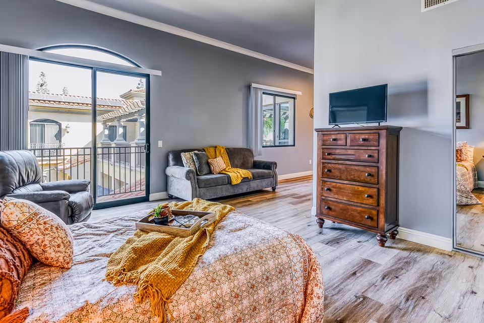 A bright bedroom with a large bed covered in patterned bedding and a mustard yellow throw blanket. There is a gray armchair and a gray sofa with cushions and a yellow throw blanket against a gray wall. A wooden dresser with a flat-screen TV on top stands near a mirrored closet door. Sliding glass doors open to a balcony with a view of a neighboring building with a tiled roof.