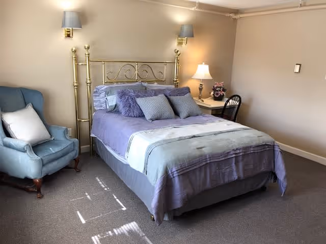 A bedroom with a brass bed frame, a bed made with purple and gray bedding, and several pillows. To the left of the bed is a blue upholstered armchair with a white pillow. On the right side of the bed is a small white nightstand with a lamp and a small flower arrangement. The walls are beige, and there are two wall-mounted lamps above the bed.