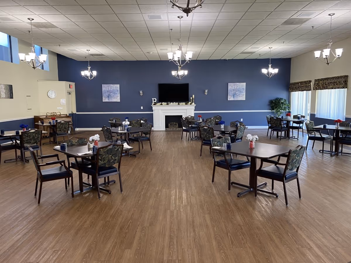 A spacious dining room with multiple tables and chairs arranged neatly on a wooden floor. The walls are painted blue and beige, with a white fireplace and a mounted TV in the center. There are chandeliers hanging from the ceiling and windows with blinds on the right side. Small flower vases are placed on each table.