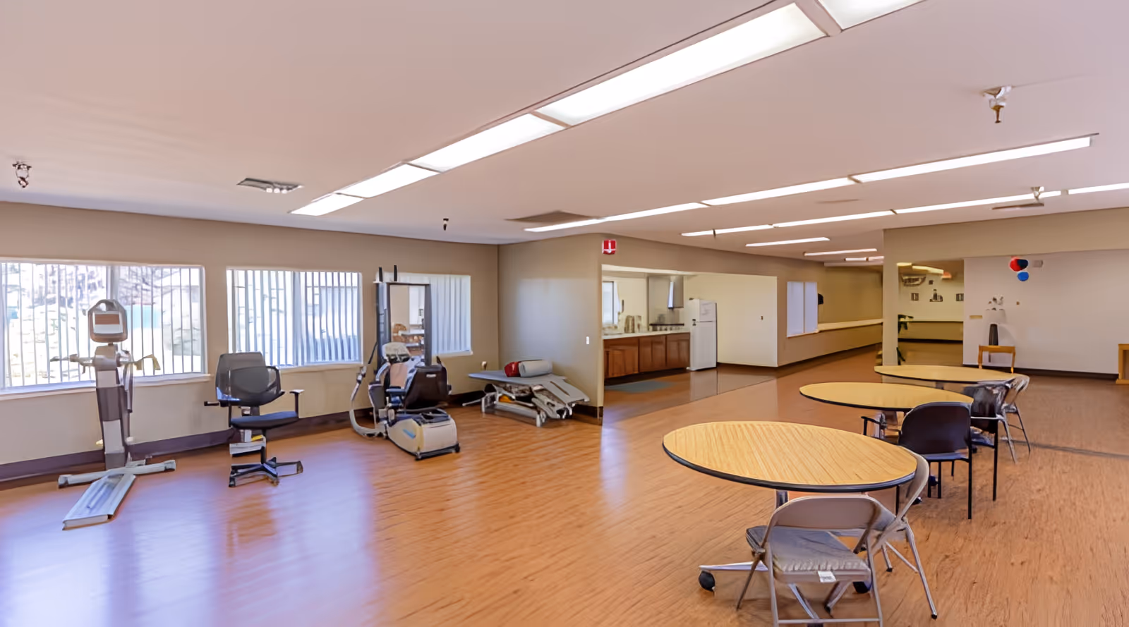 A spacious room with exercise equipment including a stationary bike and a rowing machine on the left side, and round tables with chairs on the right side. The room has large windows with bars, wood flooring, and fluorescent ceiling lights. There is a kitchenette area with a refrigerator and cabinets in the background.
