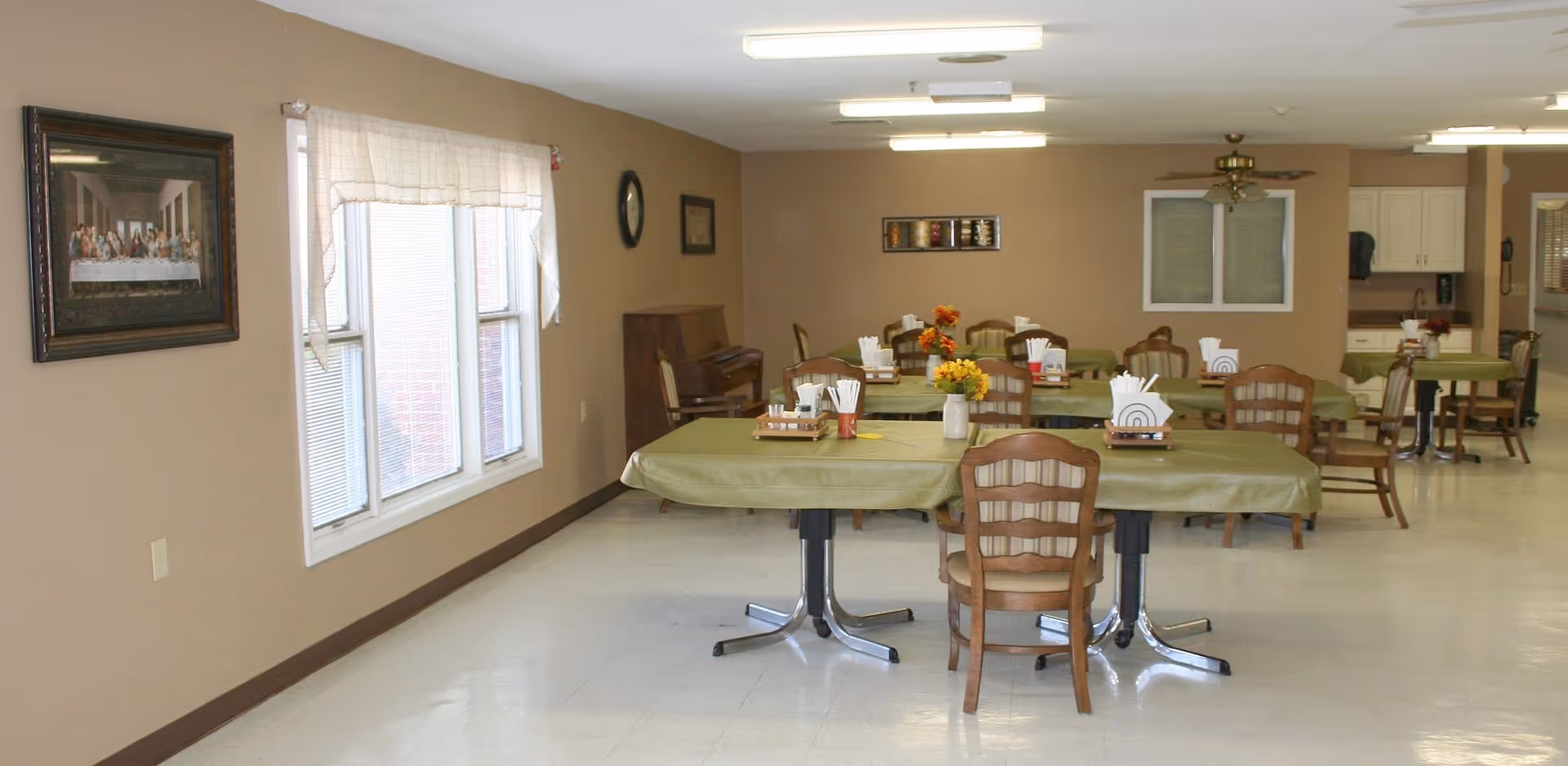 A senior living facility dining room with multiple tables covered in green tablecloths, wooden chairs, and floral centerpieces.