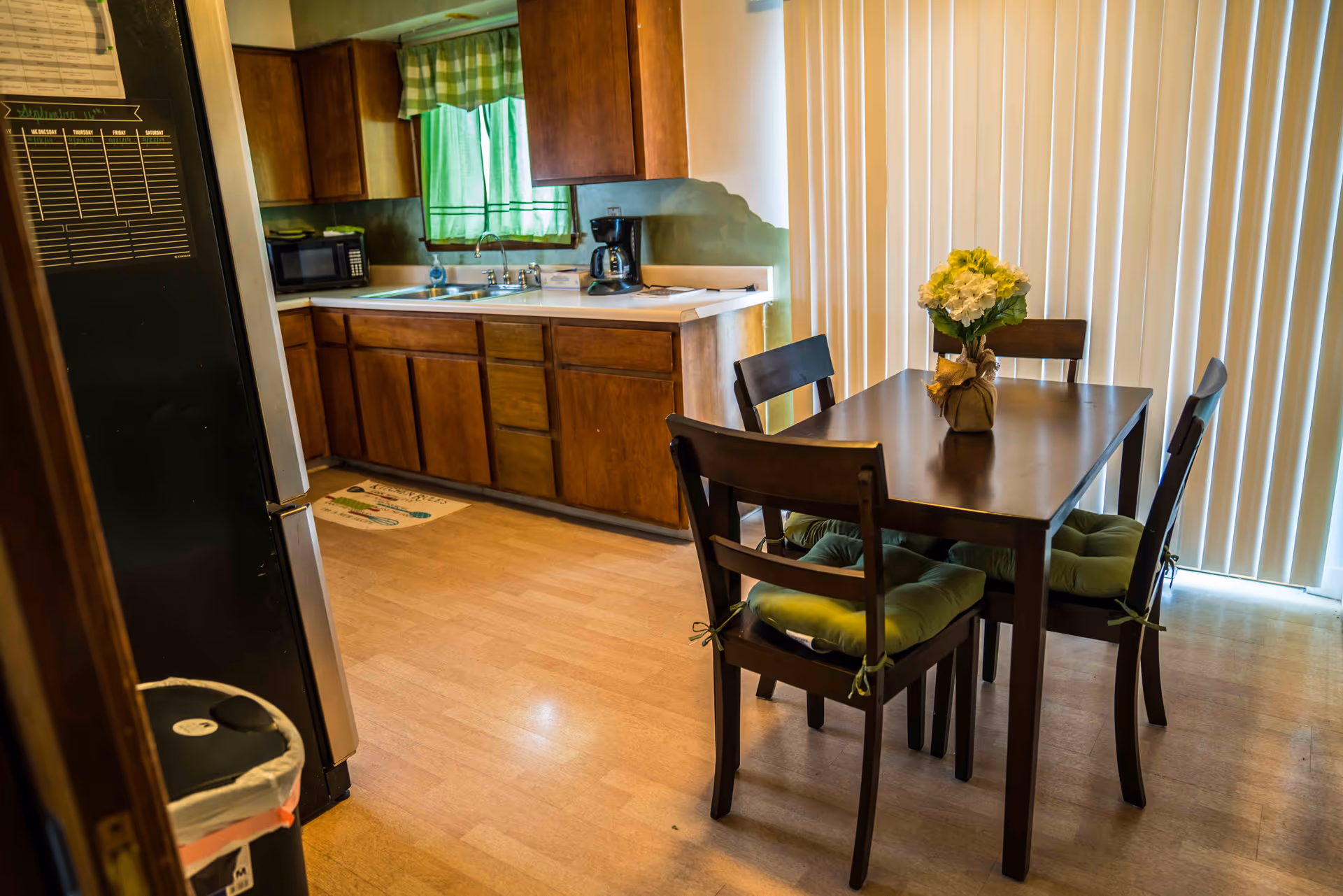 A compact kitchen and dining area with wooden cabinets, a dark wood table and four chairs with green cushions, and vertical blinds in the background.