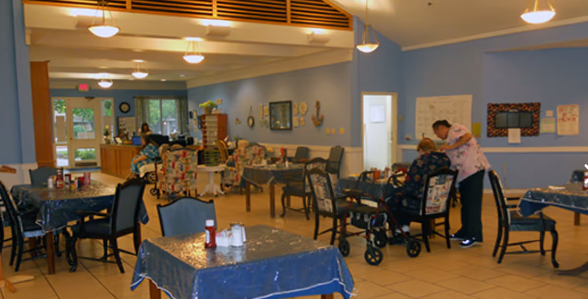 Dining/activity room with tables covered in plastic, chairs, a few residents and a caregiver assisting one resident.