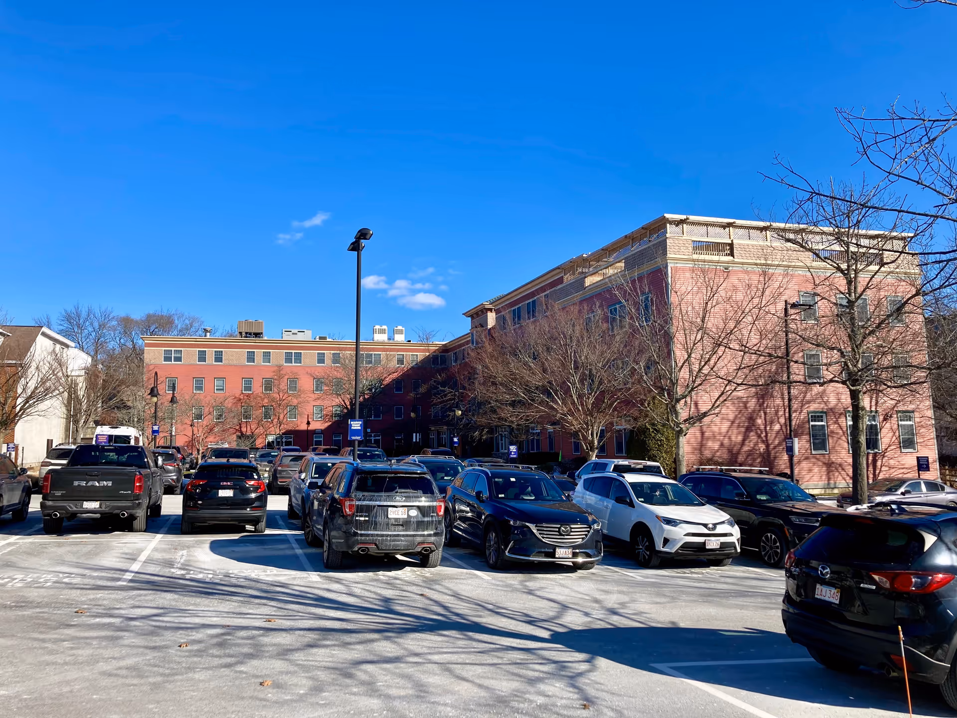 Parking lot full of cars in front of a large red-brick multi-story building under a clear blue sky.