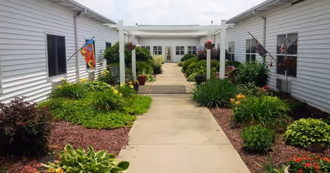 A paved walkway between two white buildings with windows, lined with green shrubs, flowering plants, and mulch beds. A white pergola structure spans the walkway, and hanging flower pots are suspended from it. Flags are displayed on the buildings, and the sky is partly cloudy.