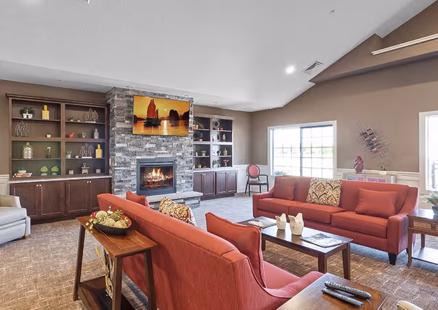 A spacious living room with two red sofas facing each other, a wooden coffee table with decorative items, a stone fireplace with a TV mounted above it, built-in wooden shelves with various decorations, large windows letting in natural light, and neutral-colored walls and carpet.