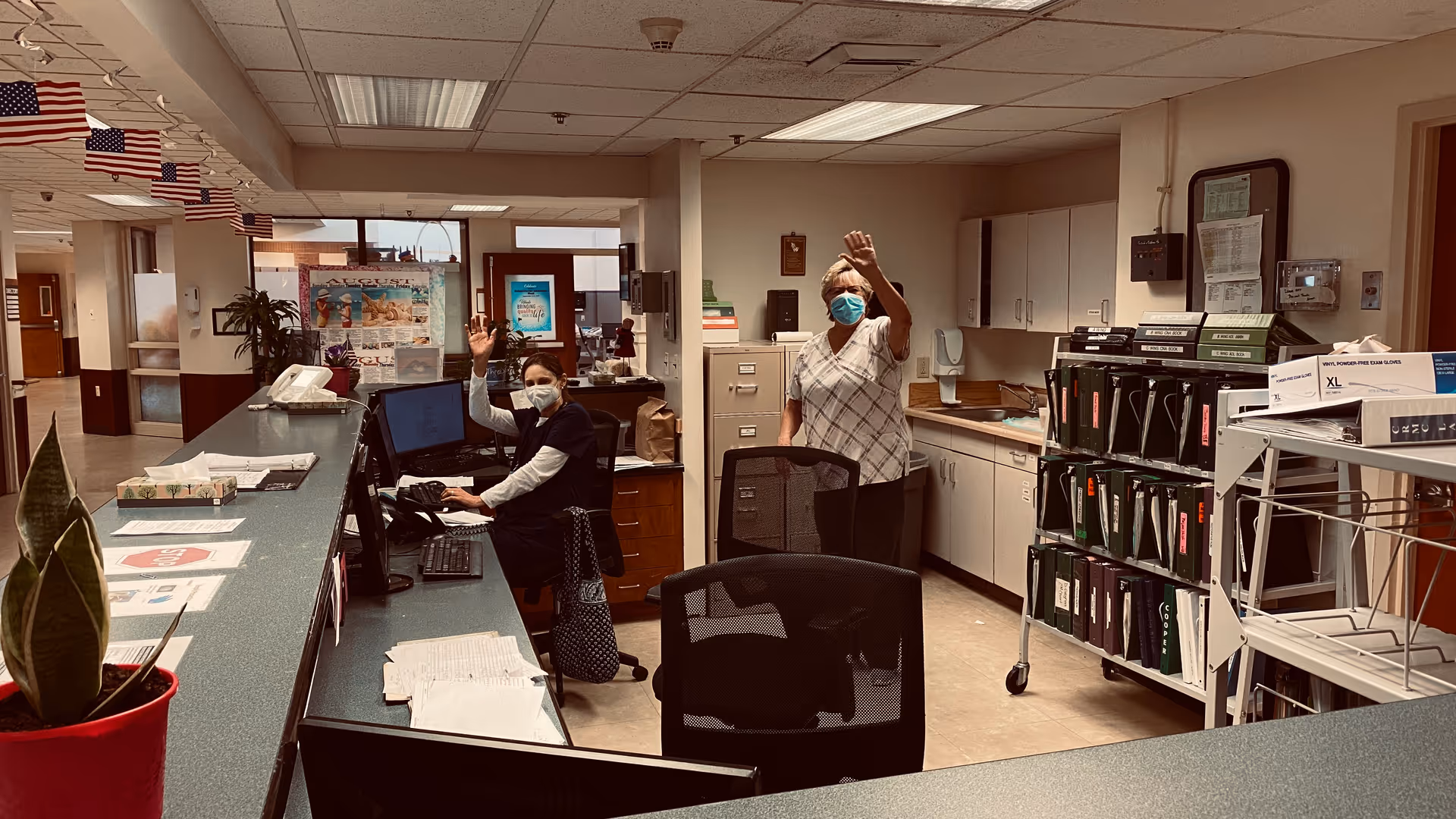 Two healthcare workers wearing masks are waving from behind a reception desk in a senior healthcare facility. The area has office furniture, filing cabinets, and medical supplies, with small American flags hanging from the ceiling.