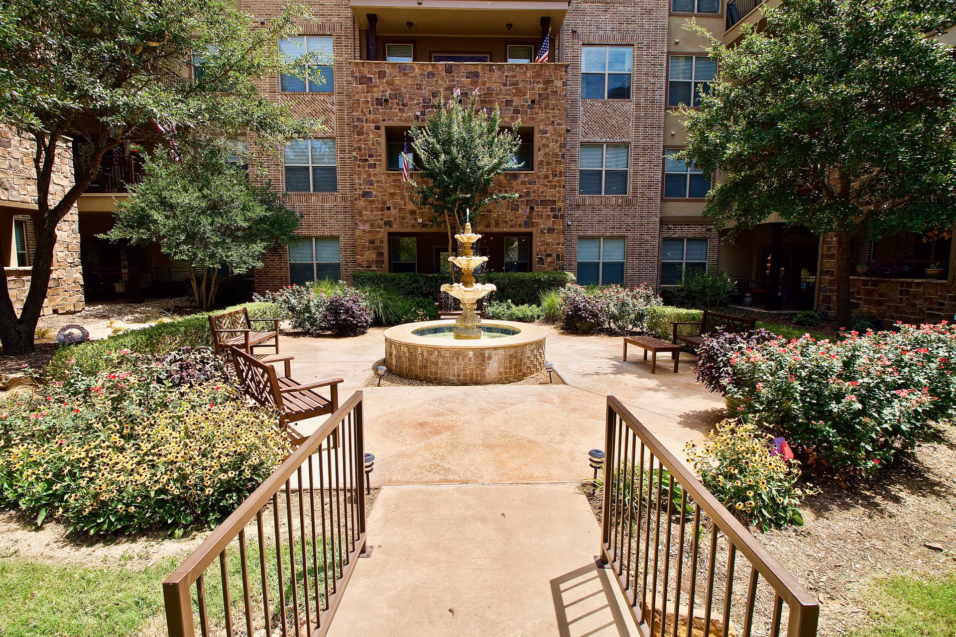 Outdoor courtyard area with a central three-tiered water fountain surrounded by benches, flowering plants, and trees, in front of a multi-story brick and stone building with windows and balconies.