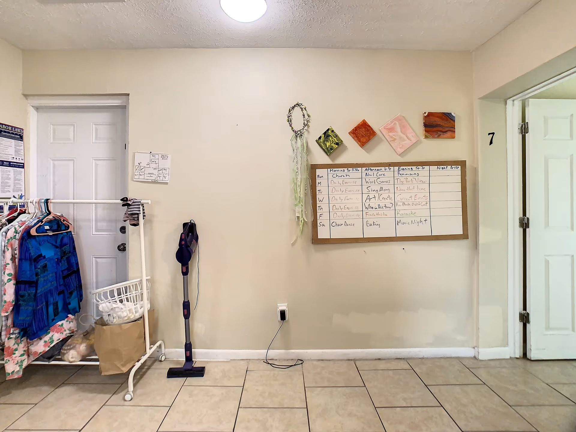 Interior view of a room with a white door on the left, a clothing rack with clothes and bags, a cordless vacuum cleaner mounted on the wall, and a whiteboard with a weekly activity schedule. The wall also has some small decorative items and a number 7 near an open door on the right.