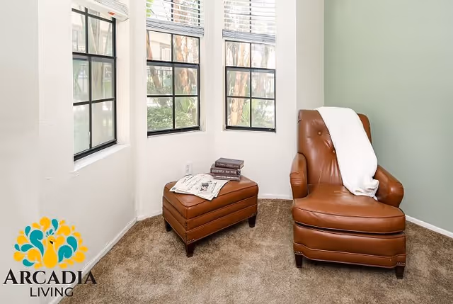 A cozy corner with a brown leather armchair draped with a white blanket, next to a matching ottoman holding two books and a newspaper. Three windows with black frames let in natural light, and the floor is carpeted in a neutral tone.