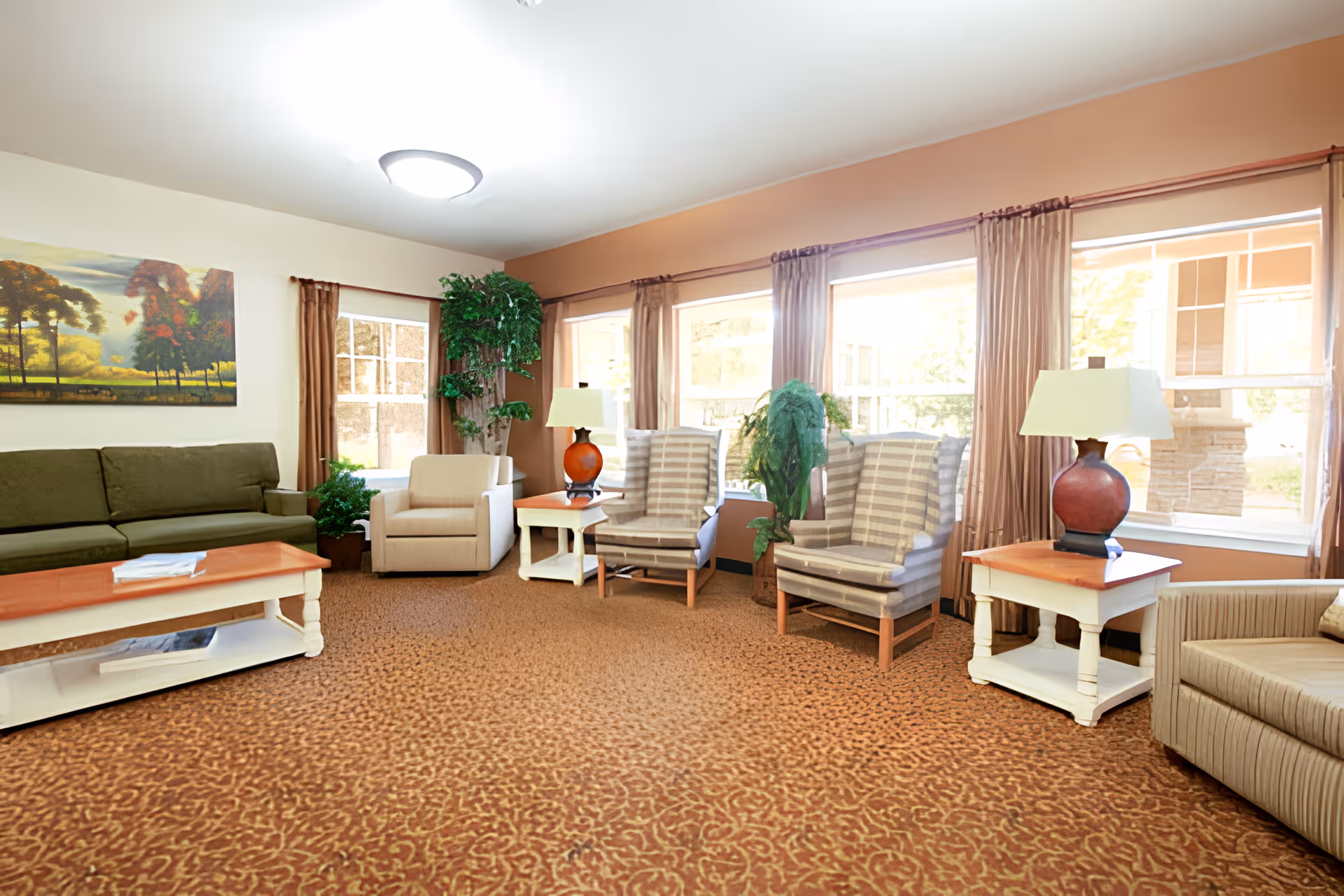 A bright and cozy senior living facility lounge with multiple seating options including a green sofa, beige armchair, two striped armchairs, and a beige couch. The room features large windows with brown curtains letting in natural light, two side tables with matching lamps, a coffee table, several potted plants, and a landscape painting on the wall.