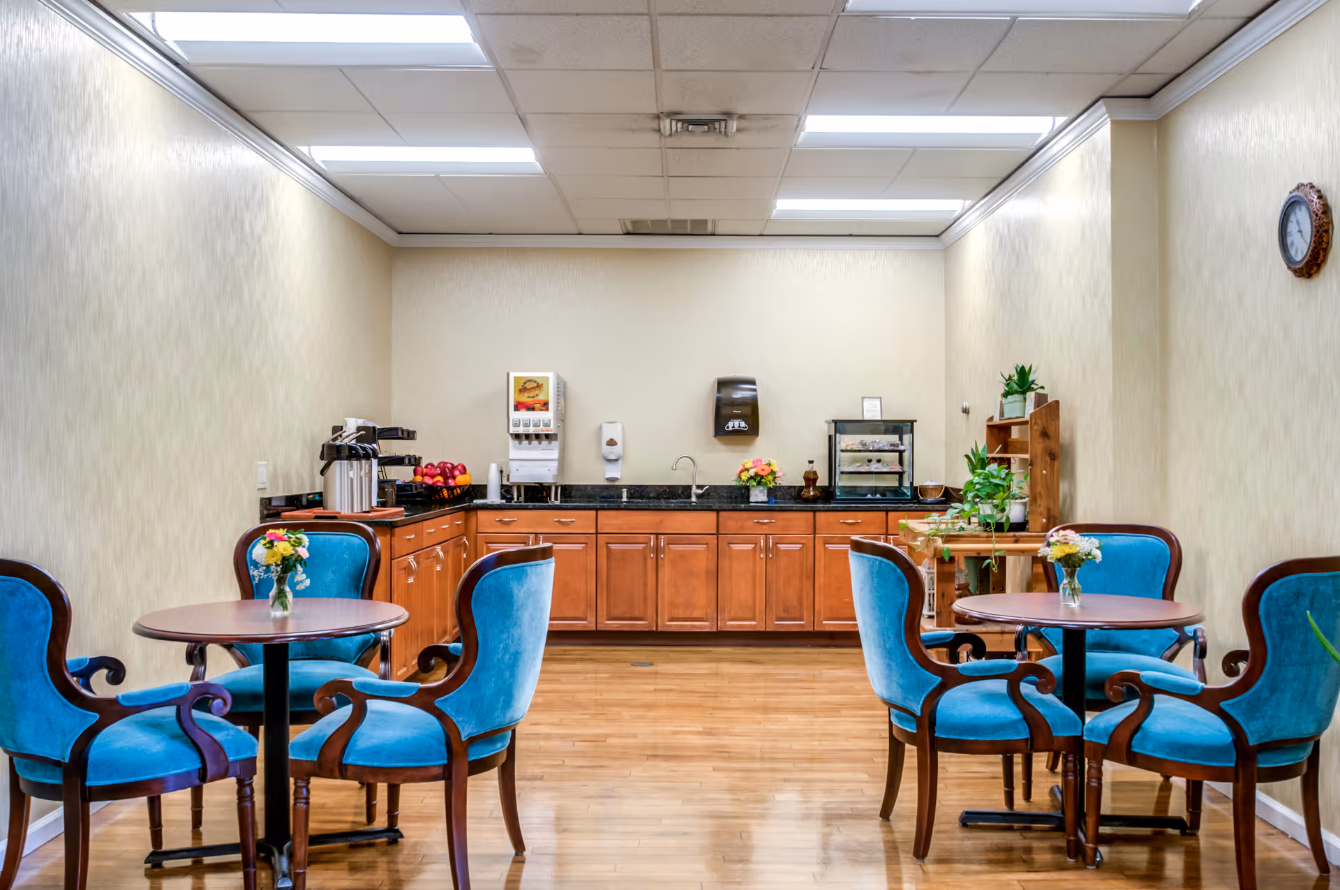 A small dining area with two round wooden tables, each surrounded by four blue upholstered chairs with wooden frames. On each table is a small vase with flowers. The back wall features a countertop with wooden cabinets underneath, a beverage dispenser, coffee urns, a hand sanitizer dispenser, and a small display case. The room has light-colored walls, a wooden floor, and a clock on the right wall.