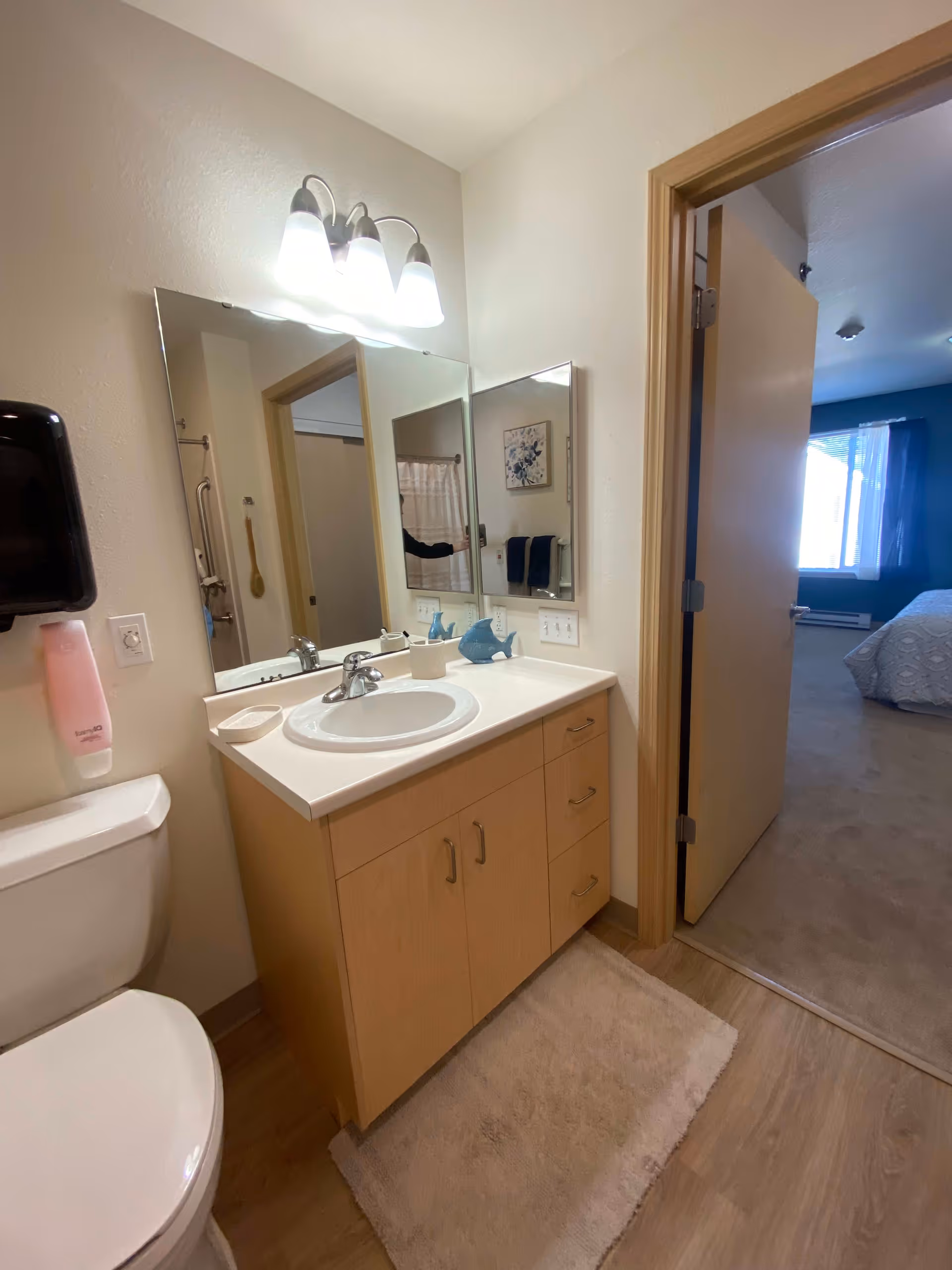 A bathroom with a white toilet, a wooden vanity with a white sink, a large mirror above the sink, and a three-light fixture. A soap dispenser is mounted on the wall next to the toilet. The bathroom door is open, revealing a carpeted bedroom with a bed and a window with curtains.