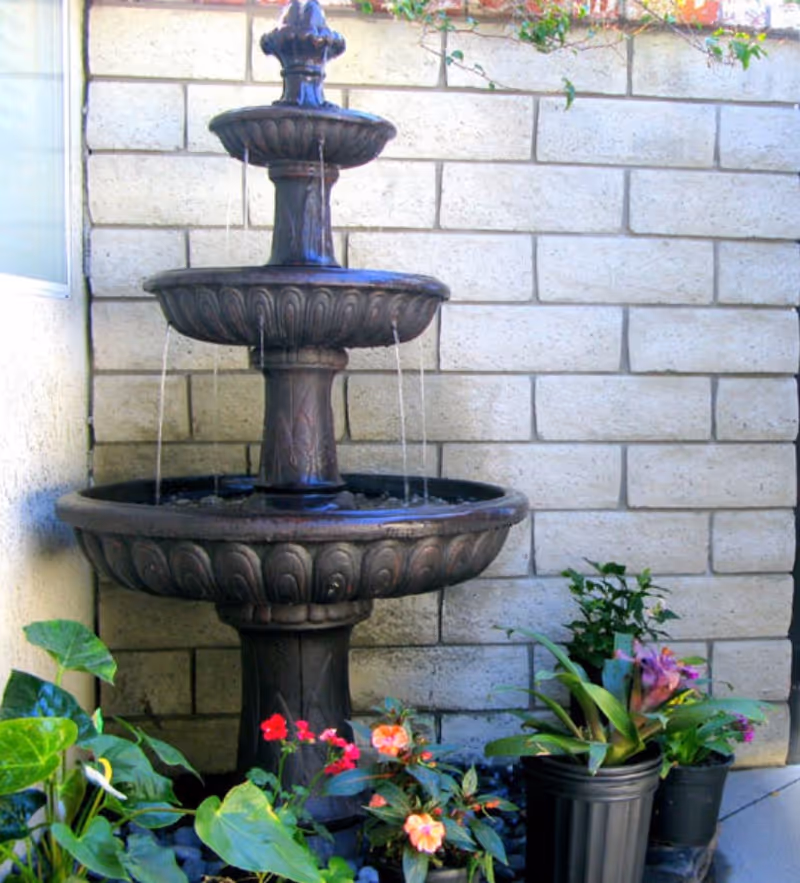 A three-tiered decorative water fountain with water flowing from the top tier to the bottom, surrounded by various potted plants with green leaves and colorful flowers, set against a light-colored brick wall.