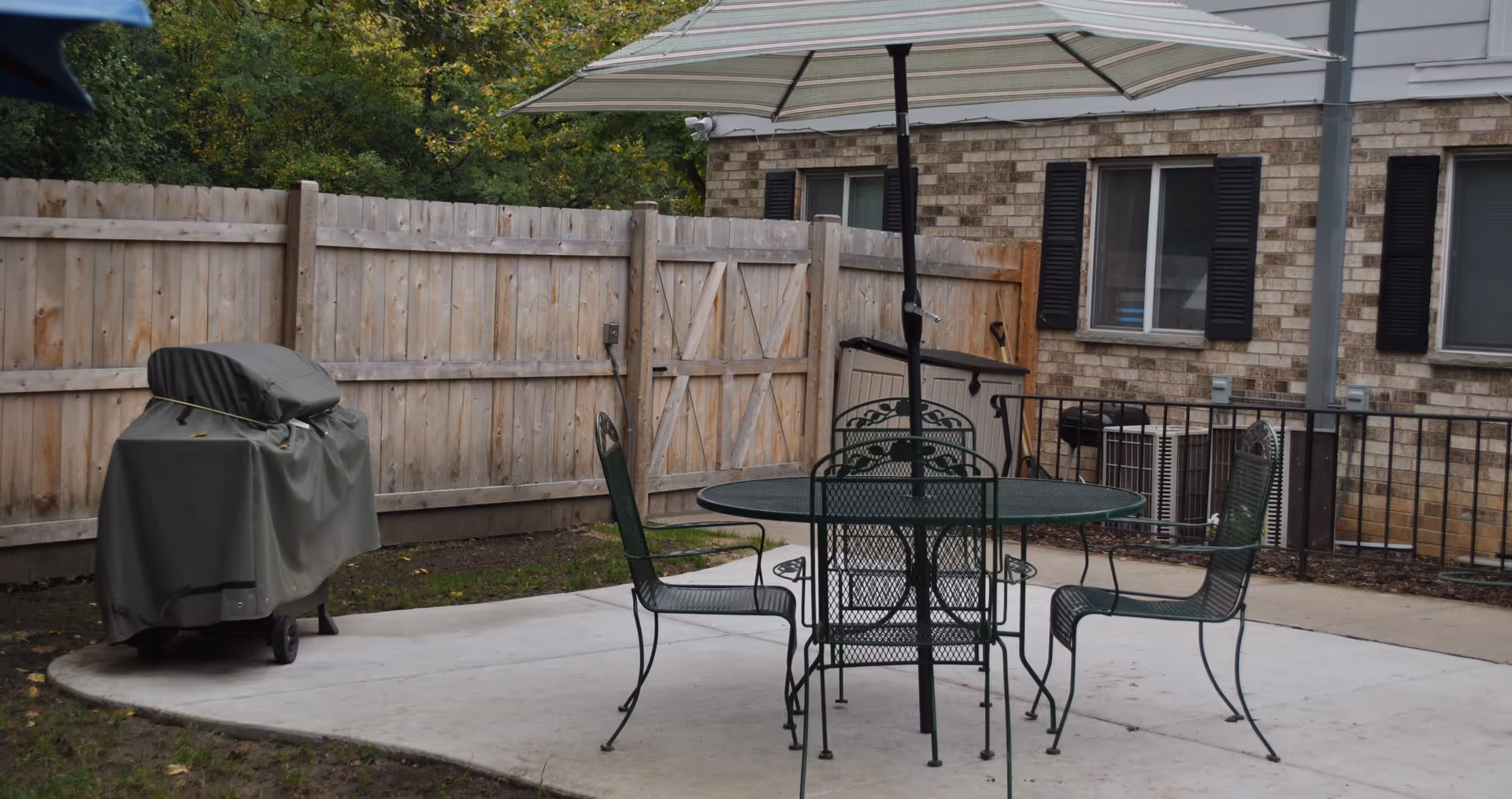 Outdoor patio area with a round metal table and four matching chairs under a large umbrella. A covered grill is positioned near a wooden fence, and a brick building with windows and black shutters is visible in the background.