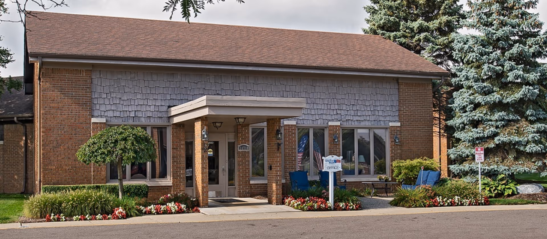 Exterior view of a single-story brick building with a brown shingled roof and a covered entrance supported by brick columns. There are windows on either side of the entrance, with American flags visible inside. The landscaping includes a small tree, bushes, colorful flowers, and a large evergreen tree to the right. A sign near the entrance reads 'American House East II Office'.