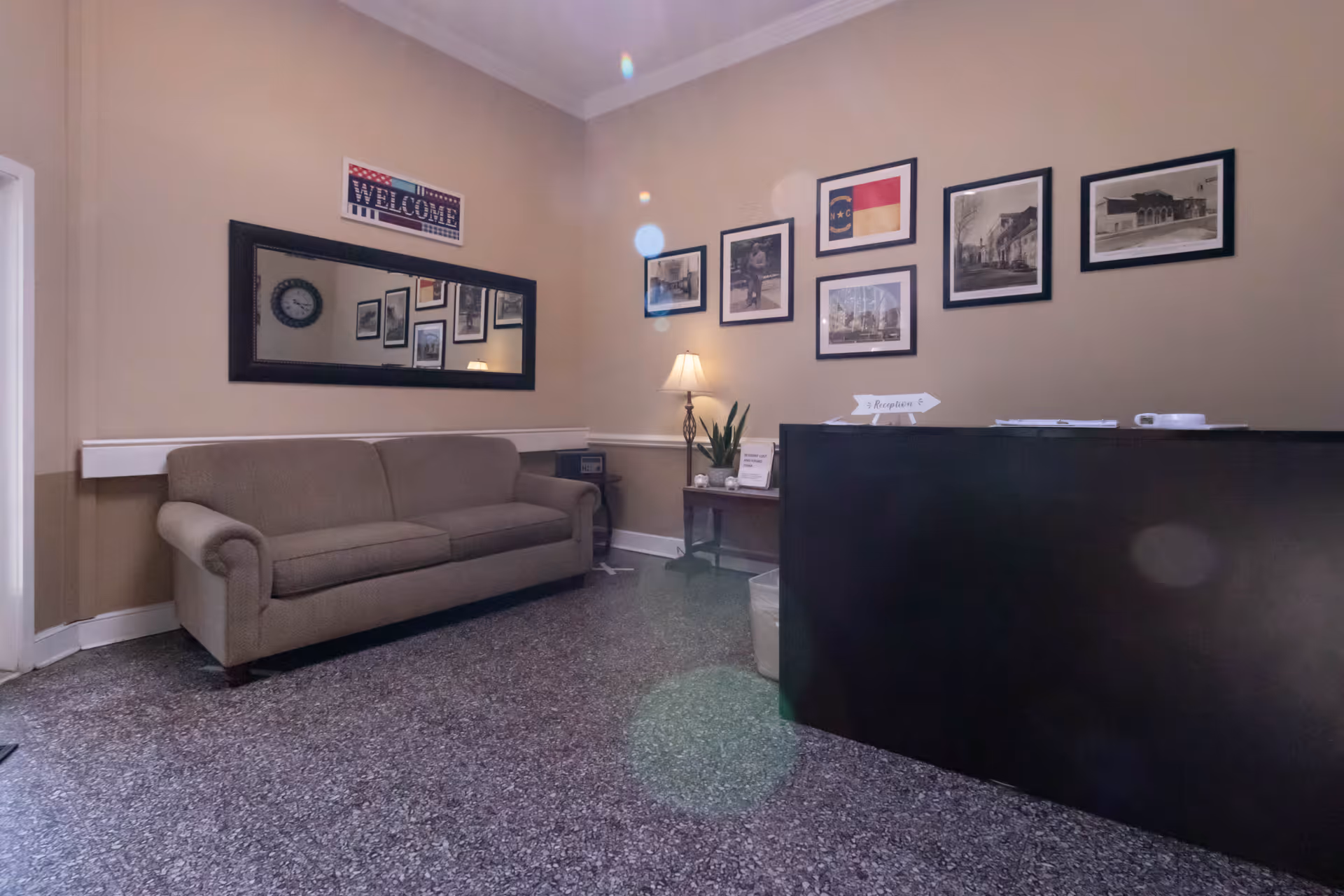 Reception area with a brown sofa against the left wall, a large rectangular mirror above it, and a small table with a lamp and plant in the corner. Several framed pictures and a welcome sign are hung on the beige walls. A black reception desk is on the right side with papers and a small sign on top.