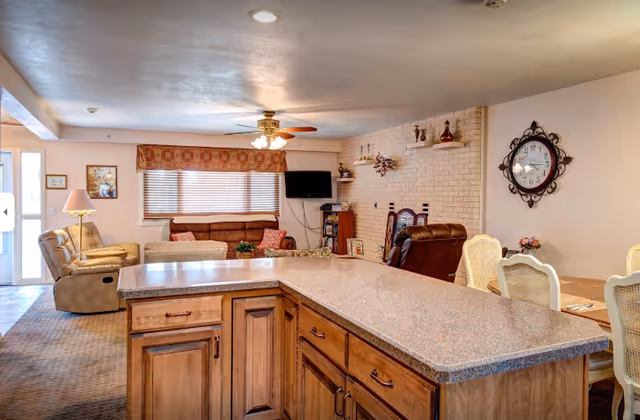 Open-plan living area with a kitchen island in the foreground, sofa and recliners, wall-mounted TV, and dining table in the background.